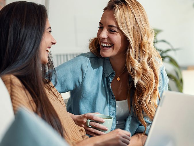 Due donne sorridenti sedute su un divano davanti a un computer portatile